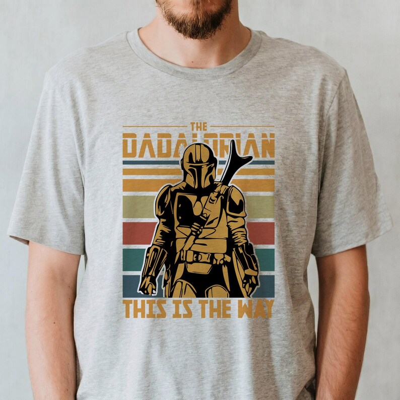 May include: A light grey t-shirt with a graphic of a Mandalorian-style character in gold and black armour, with a weapon on their back. The text "THE DADALORIAN" and "THIS IS THE WAY" are printed above and below the character, respectively, in a retro style.