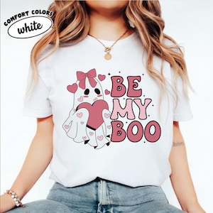 May include: A white t-shirt featuring a graphic of a ghost with a pink bow holding a large pink heart, surrounded by smaller pink hearts and the text "BE MY BOO" in a bold, retro-style font. The shirt has short sleeves and a crew neckline.