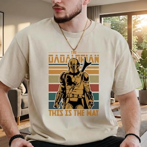 May include: Beige t-shirt with a graphic of The Mandalorian. The design includes the text "The Dadalorian" above an illustration of the character, with the phrase "This is the Way" below. The design incorporates a colour scheme of brown, orange, red, green, and blue.