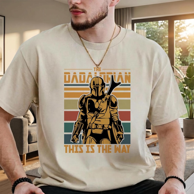 May include: Beige t-shirt with a graphic of The Mandalorian. The design includes the text "The Dadalorian" above an illustration of the character, with the phrase "This is the Way" below. The design incorporates a colour scheme of brown, orange, red, green, and blue.