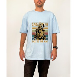 May include: Pale blue t-shirt with a retro graphic of a Mandalorian warrior. The design includes the text "THE DADALORIAN THIS IS THE WAY" and a helmeted figure, set against brown, green, and blue horizontal stripes.
