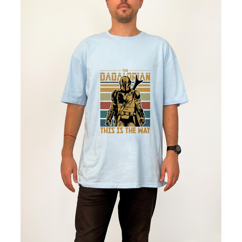 May include: Pale blue t-shirt with a retro graphic of a Mandalorian warrior. The design includes the text "THE DADALORIAN THIS IS THE WAY" and a helmeted figure, set against brown, green, and blue horizontal stripes.