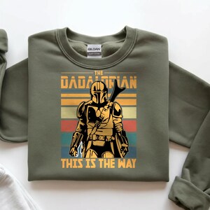 May include: Khaki green sweatshirt with a retro graphic of a Mandalorian character. The design includes the text "THE DADALORIAN" and "THIS IS THE WAY". The image shows a figure in armour holding a weapon, set against horizontal stripes.