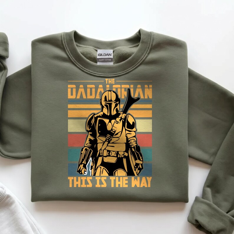 May include: Khaki green sweatshirt with a retro graphic of a Mandalorian character. The design includes the text "THE DADALORIAN" and "THIS IS THE WAY". The image shows a figure in armour holding a weapon, set against horizontal stripes.
