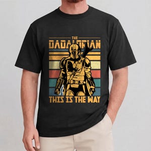 May include: Black t-shirt with a graphic of a Mandalorian warrior and the text "THE DADALORIAN" above and "THIS IS THE WAY" below. The design has a retro colour scheme with horizontal stripes in orange, yellow, red, teal, and brown.