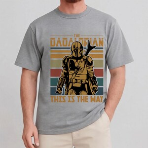 May include: Grey t-shirt with a graphic of a character from "The Mandalorian", with the text "THE MANDALORIAN" and "THIS IS THE WAY" in a retro style. The design includes a figure in armour and a striped background.
