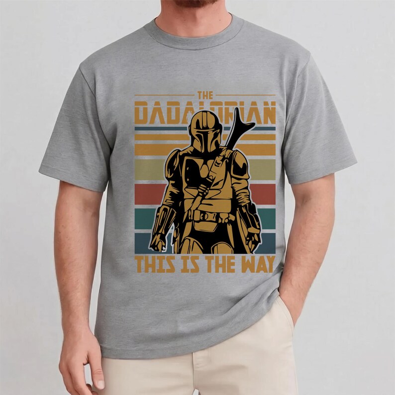 May include: Grey t-shirt with a graphic of a character from "The Mandalorian", with the text "THE MANDALORIAN" and "THIS IS THE WAY" in a retro style. The design includes a figure in armour and a striped background.