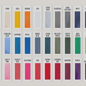 May include: A display of colour swatches, each labelled with a colour name. Colours include white, sand, gold, orange, ash, sport grey, charcoal, dark heather, navy, and black. Additional colours are light blue, Carolina blue, sapphire, royal, indigo blue, legion blue, military green, forest green, irish green, and kiwi.
