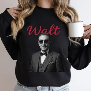 Close-up of a woman holding a white enamel mug while wearing a black retro Disneyland sweatshirt. The apparel shows a vintage Walt Disney graphic with striking red cursive lettering.