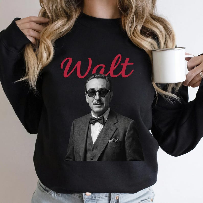 Close-up of a woman holding a white enamel mug while wearing a black retro Disneyland sweatshirt. The apparel shows a vintage Walt Disney graphic with striking red cursive lettering.