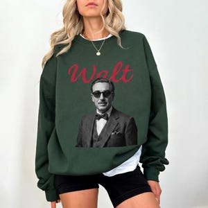 A woman modeling a dark green Magic Kingdom sweatshirt. The design features a retro black and white portrait of Walt Disney in a suit and sunglasses, with "Walt" written in red.