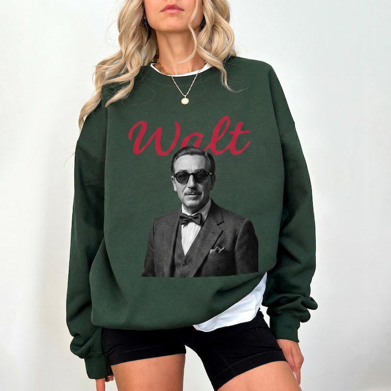 A woman modeling a dark green Magic Kingdom sweatshirt. The design features a retro black and white portrait of Walt Disney in a suit and sunglasses, with "Walt" written in red.