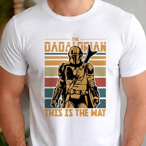May include: White t-shirt with a graphic of The Mandalorian. The design features a gold-coloured Mandalorian figure with a weapon, set against a backdrop of horizontal stripes in brown, orange, teal, and blue. The text "THE MANDALORIAN" and "THIS IS THE WAY" are displayed.