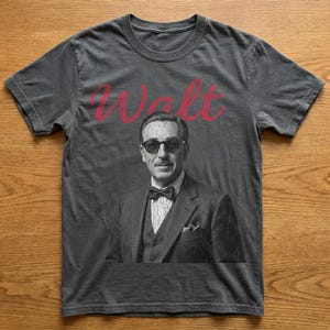 Dark gray vintage-wash magic kingdom tee laid on a wooden background, featuring a distressed, cracked-effect black and white portrait of Walt Disney in sunglasses with red text above.