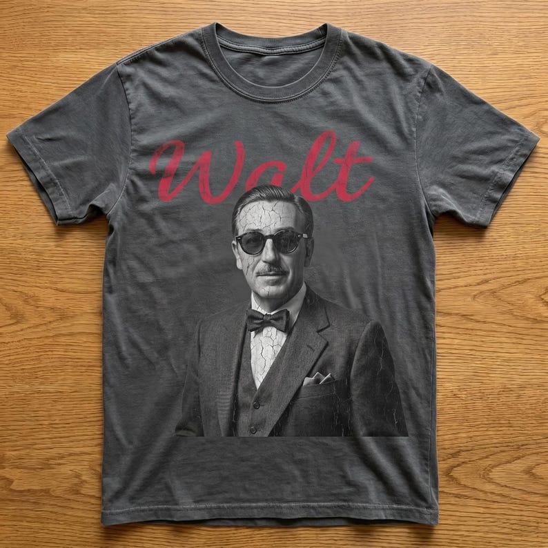 Dark gray vintage-wash magic kingdom tee laid on a wooden background, featuring a distressed, cracked-effect black and white portrait of Walt Disney in sunglasses with red text above.