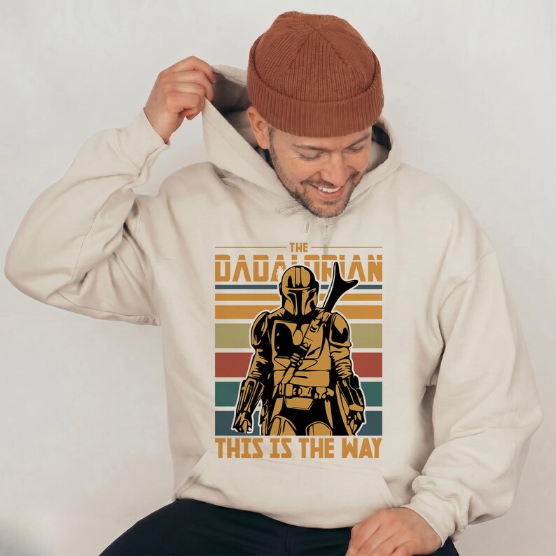 May include: Beige hoodie with a vintage-style graphic of The Mandalorian. The design includes a helmeted figure, the text "THE MANDALORIAN", and "THIS IS THE WAY", set against horizontal stripes in various colours.