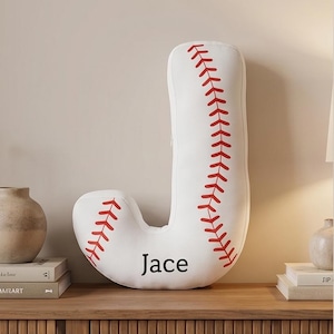 May include: A white baseball-themed letter "J" pillow with red stitching and the name "Jace" printed in black. The pillow is on a wooden surface with books and decorative items in the background.