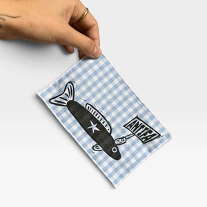 May include: A rectangular fabric patch featuring a light blue and white gingham pattern. A black fish illustration is printed on the fabric, holding a small sign that reads ANTIFA in black capital letters. The fish features a white star.