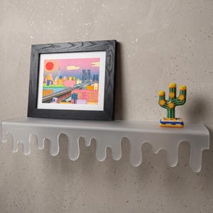 May include: A clear, dripping-edge shelf holds a framed print of a city skyline with a red sun. A small, crocheted cactus with yellow flowers sits to the right. The frame is black, and the shelf is mounted on a textured wall.