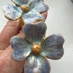 May include: Two flower-shaped drawer knobs featuring four petals each with a swirling blend of light blue, lavender, and white enamel. The petals are outlined in gold-toned metal, and each center features a textured, rounded gold-toned stamen.