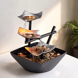 May include: A three-tiered indoor water fountain with a black base and frame. The fountain features three silver-colored, leaf-shaped bowls, each filled with stones. Water cascades from the top bowl to the bottom, creating a tranquil ambiance.