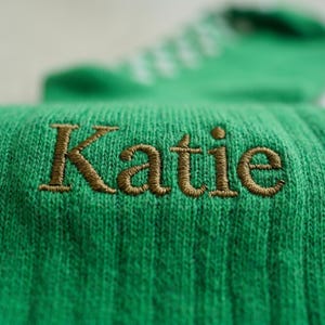 Op de afbeelding: Close-up van een groene sok met de naam "Katie" geborduurd in bruin garen. De sok heeft een geribbelde textuur en is gemaakt van een zacht, gebreid materiaal. De achtergrond is wazig, waardoor de details van de sok worden benadrukt.