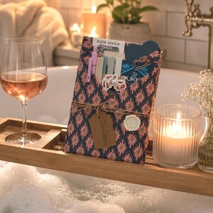 May include: A bamboo bathtub tray holds a glass of rose wine, a gift-wrapped book with a floral pattern, a brown paper tag labeled Thriller, a white wax seal, pens, and stickers. A glass candle and a small vase of baby's breath flowers are also present.