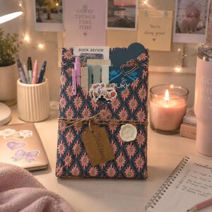 May include: A rectangular book sleeve featuring a dark blue fabric with a repeating pink and green floral diamond pattern. The sleeve is wrapped with twine, a brown paper tag, and a white wax seal, containing pens, bookmarks, and stickers.
