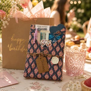 May include: A rectangular gift package wrapped in dark blue paper with a pink floral pattern, tied with twine and a brown tag labeled Thriller. The package contains stationery items, including pens and cards with the text Book Review and JURK.