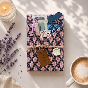 May include: A rectangular package wrapped in dark blue paper with a pink floral diamond pattern, tied with twine. It contains a book review card, pens, sticky notes, stickers, and a metal clip. A brown tag labeled Thriller and a wax seal are attached.