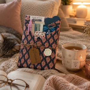 May include: A rectangular book sleeve featuring a dark blue and pink floral diamond pattern, tied with twine and a brown tag labeled Thriller. The sleeve holds pens, sticky notes, and a blue card. A mug with a tea bag tag reading you've got this sits nearby.