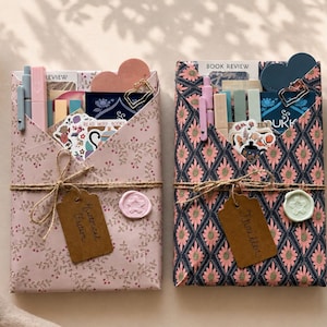 May include: Two rectangular gift-wrapped packages featuring stationery items tucked into the top folds. The left package has light pink floral paper, a brown tag labeled Historical Fiction, and a pink wax seal. The right package has a dark blue and pink geometric pattern, a brown tag labeled Thriller, and a light green wax seal.