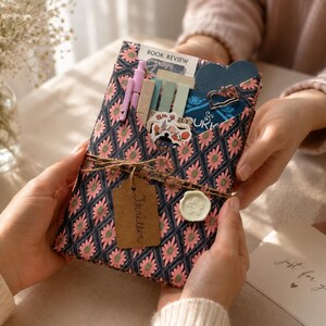 May include: A rectangular fabric pouch featuring a dark blue diamond pattern with pink flowers. The pouch contains a light pink pen, several pastel sticky note tabs, a blue heart-shaped card, and a sticker. It is tied with twine and a brown tag labeled Thriller.