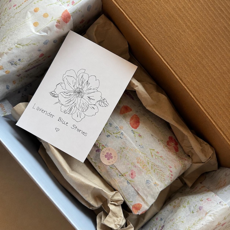 May include: A cardboard shipping box containing items wrapped in floral tissue paper and a white card with a black line drawing of a flower and the text Lavender Blue Stories with a small heart symbol printed below the text.