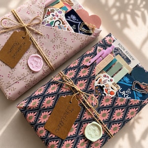 May include: Two gift-wrapped packages featuring floral patterns, tied with twine and brown paper tags labeled Historical Fiction and Thriller. Each includes a wax seal, a pen, colorful sticky notes, stickers, a heart-shaped cutout, and a metal book-shaped paperclip.