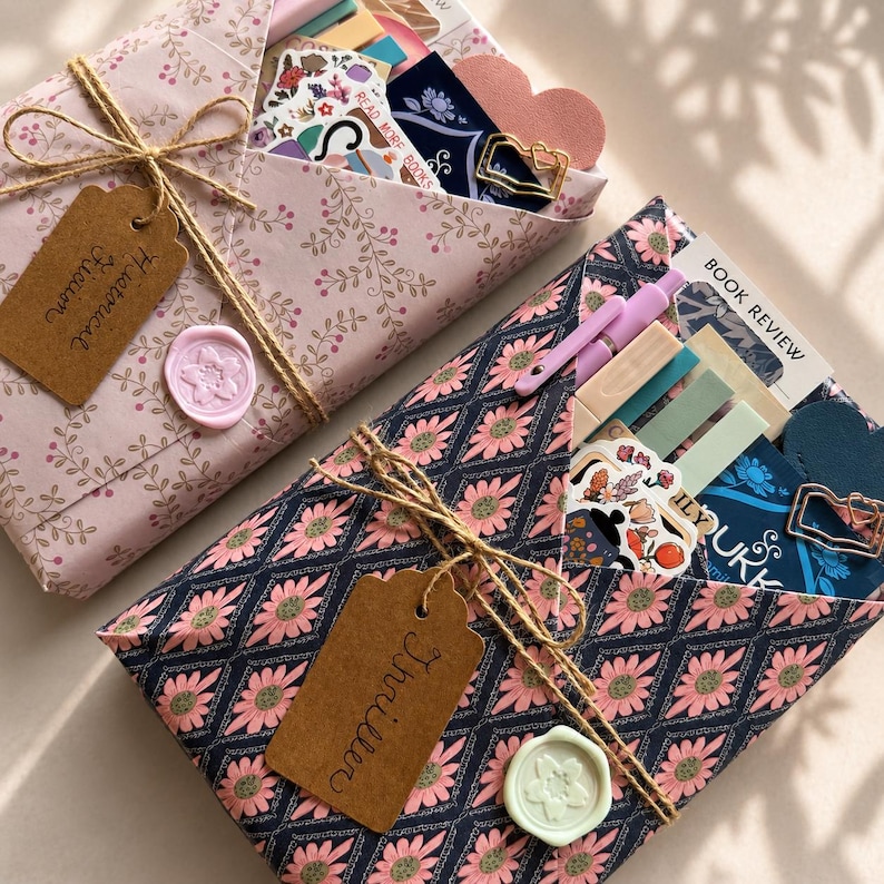 May include: Two gift-wrapped packages featuring floral patterns, tied with twine and brown paper tags labeled Historical Fiction and Thriller. Each includes a wax seal, a pen, colorful sticky notes, stickers, a heart-shaped cutout, and a metal book-shaped paperclip.