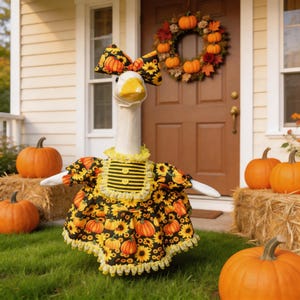 May include: A white goose figurine wearing a black dress with a yellow sunflower and orange pumpkin print, featuring a matching bow, a yellow ruffled collar, and two yellow buttons on the bodice, standing on a lawn with orange pumpkins.