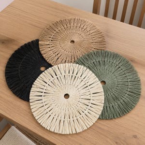 Op de afbeelding: Een set van vier ronde gevlochten placemats met een radiaal patroon en een gat in het midden. De placemats zijn gemaakt van vezelig materiaal in vier verschillende kleuren: natuurlijk beige, zwart, gebroken wit en gedempt saliegroen.
