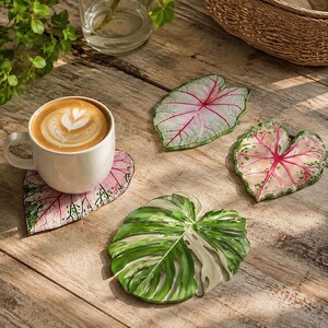Könnte beinhalten: Eine weiße Keramiktasse mit Latte Art steht auf einem herzförmigen Untersetzer mit rosa-grünem Blattmuster. Drei weitere blattförmige Untersetzer sind in der Nähe angeordnet, darunter zwei mit rosa-grünem Caladium-Design und einer mit grün-weißem Monstera-Blatt-Design.