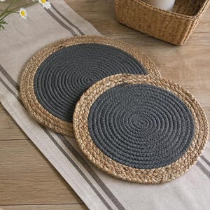 May include: Two round placemats featuring a coiled design with a dark grey centre and a natural tan braided border. The placemats are constructed from woven rope and straw-like material, showcasing a circular pattern that radiates from the centre.