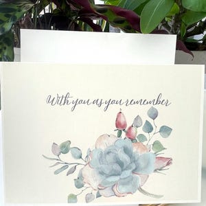May include: A cream-coloured greeting card featuring the text "With you as you remember" in dark grey script. Below the text is a watercolour illustration of a large blue-green succulent surrounded by small pink berries and sage green leaves.