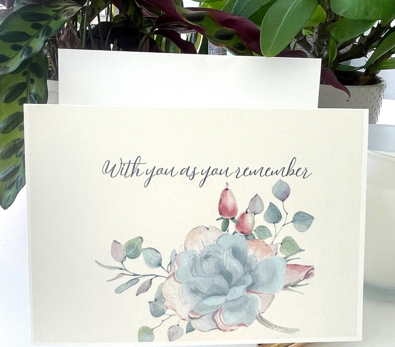 May include: A cream-coloured greeting card featuring the text "With you as you remember" in dark grey script. Below the text is a watercolour illustration of a large blue-green succulent surrounded by small pink berries and sage green leaves.