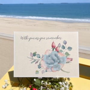 May include: A rectangular greeting card with a cream-coloured background featuring a watercolour illustration of a large blue-green succulent surrounded by pink buds and green leaves. The text "With you as you remember" is printed in black script.