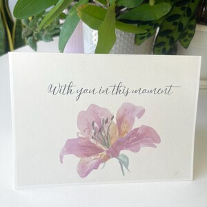 May include: A rectangular off-white greeting card featuring a watercolour illustration of a pink lily flower centred at the bottom. Above the flower, the text "With you in this moment" is printed in a dark grey, elegant cursive script font.