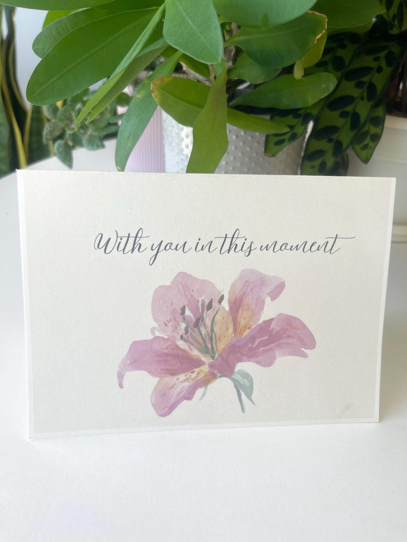 May include: A rectangular off-white greeting card featuring a watercolour illustration of a pink lily flower centred at the bottom. Above the flower, the text "With you in this moment" is printed in a dark grey, elegant cursive script font.