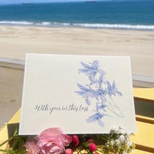 May include: A rectangular greeting card with a light cream background featuring a watercolour illustration of light blue iris flowers on the right side and the black cursive text "With you in this loss" printed on the lower left side.