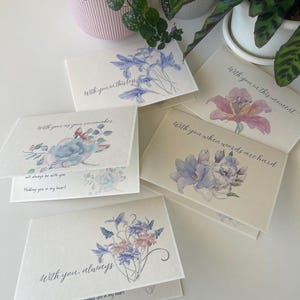 May include: A collection of folded greeting cards with floral watercolor designs in shades of blue, pink, and purple. Each card features a different floral arrangement and a handwritten message, such as "With you in this loss" and "With you, always."
