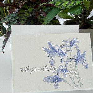 May include: A rectangular greeting card featuring a watercolour illustration of light blue iris flowers on the right side. The text "With you in this loss" is printed in a dark grey cursive script on the lower left side of the white card.