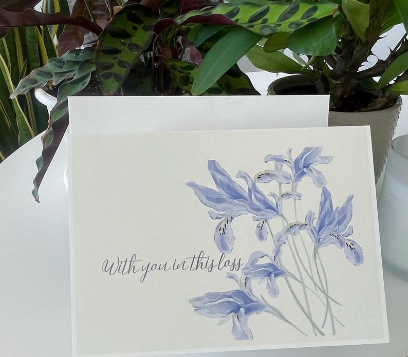 May include: A rectangular greeting card featuring a watercolour illustration of light blue iris flowers on the right side. The text "With you in this loss" is printed in a dark grey cursive script on the lower left side of the white card.