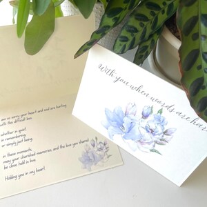 May include: A folded sympathy card with a white exterior featuring a watercolour illustration of blue flowers and the text "With you when words are hard." The interior is cream-coloured with black printed text expressing condolences and support.